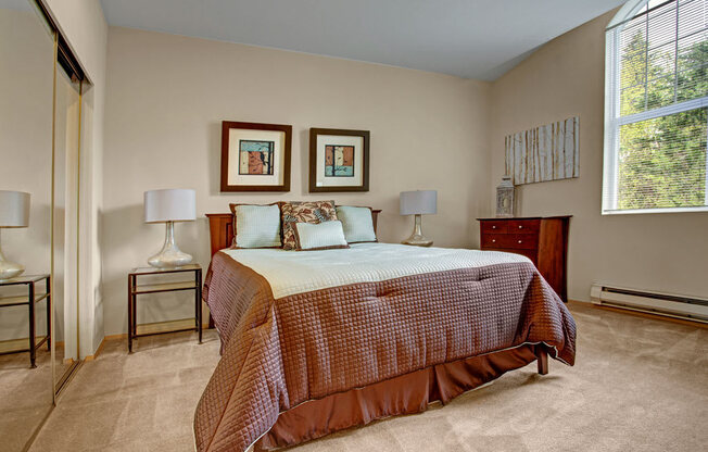 A cozy bedroom here at Aspen Creek featuring a spacious bed dressed in layered neutral bedding with accent pillows, matching bedside tables with soft table lamps, and framed wall art above the headboard, complemented by plush carpet flooring, mirrored closet doors, a wood dresser, and a large window that brings in natural light and treetop views.
