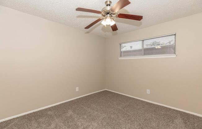 A vacant room with beige walls and carpeted flooring. A ceiling fan with light fixtures is mounted on the ceiling. A window with blinds is visible on one wall, providing natural light. The space appears neat and ready for furnishing.