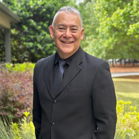 A man in a black suit and tie standing in front of a greenery.