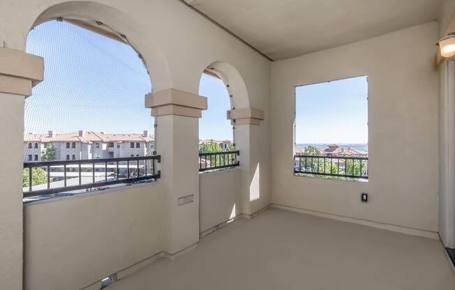 A spacious balcony featuring arched openings with screens, offering scenic views of a neighborhood with residential buildings and greenery under a clear blue sky. The floor is smooth and light-colored, creating an open and inviting atmosphere.