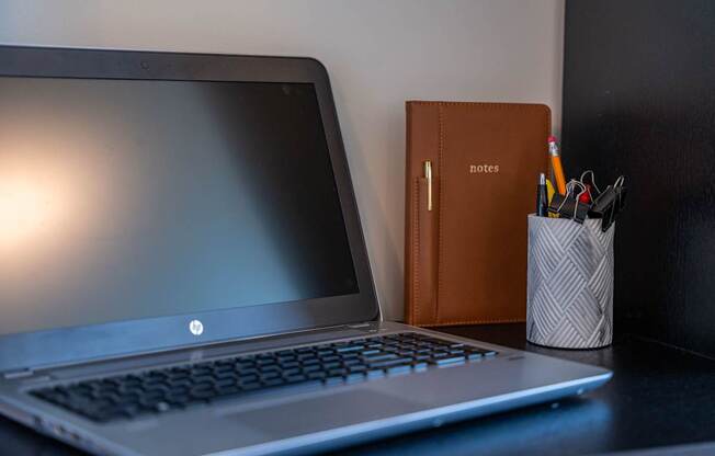 A laptop with a notebook and a cup of pens on the side on a desk at the Mastlight.