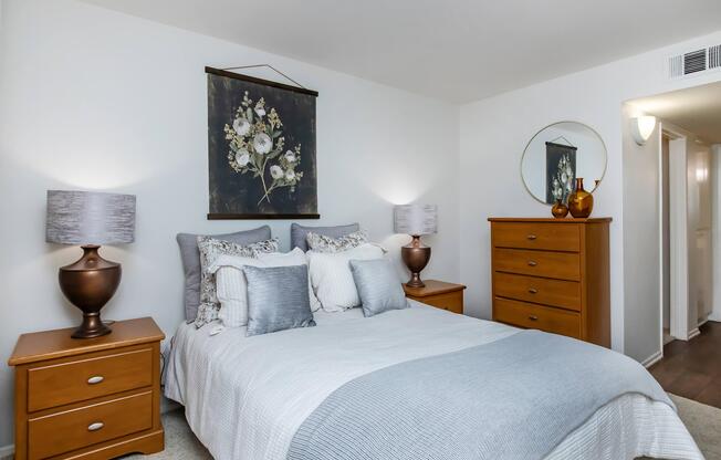 A cozy bedroom featuring a queen-sized bed with soft gray and white bedding. Two wooden bedside tables with matching lamps sit on either side. A large piece of floral artwork hangs above the bed, and a round mirror is mounted on the wall. A wooden dresser is partially visible in the background.