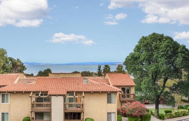 A building with a red tile roof is surrounded by greenery and a body of water.