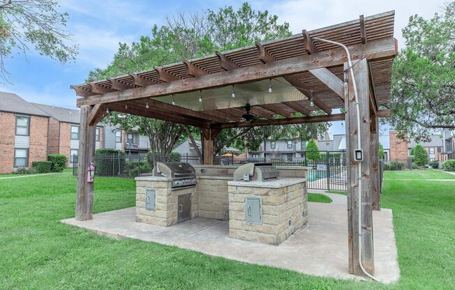 Outdoor barbecue area featuring two stone grills under a wooden pergola. The space is surrounded by grass and trees, with string lights installed for ambiance. It is located near a residential complex, providing a communal cooking space for gatherings.