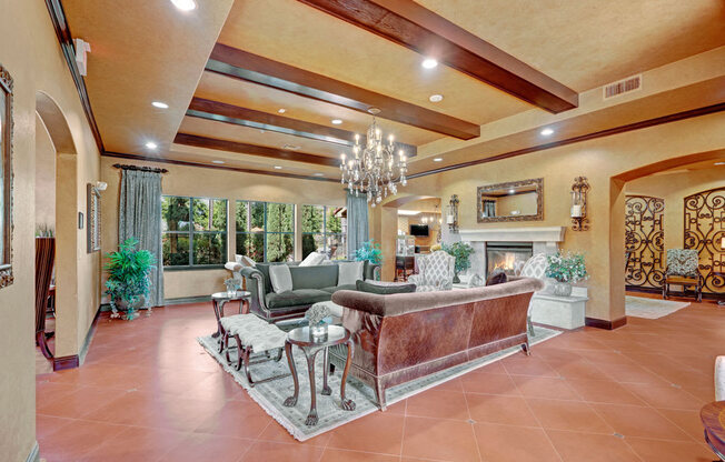 A spacious community lounge here at Tuscany at Faudree with elegant seating arranged on an area rug beneath exposed ceiling beams and a statement chandelier. Plush sofas, accent chairs, and side tables surround a decorative fireplace, while large windows, greenery, and warm finishes create an inviting space for relaxing and gathering.