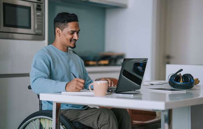 A man in a blue sweater is sitting at a table with a laptop and a cup of coffee.