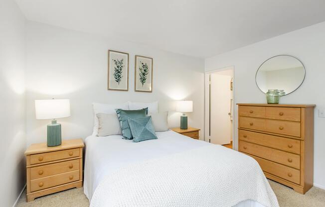 A cozy bedroom featuring a light-colored bed with decorative pillows, flanked by two bedside tables with green lamps. The room includes a wooden dresser, a round mirror, and framed botanical art on the walls, all set against a neutral backdrop, creating a warm and inviting atmosphere.