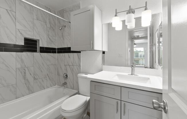 A modern bathroom featuring a white bathtub, a sleek vanity with a sink, and a large mirror. The walls are adorned with marble tiles, and thereâs a stylish light fixture above the mirror. The design is clean and contemporary, emphasizing functionality and aesthetic appeal.