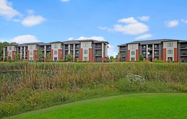 Modern three-story 49 West Apartment buildings with a mix of beige and brick exteriors sit behind a lush wetland, under a clear blue sky.