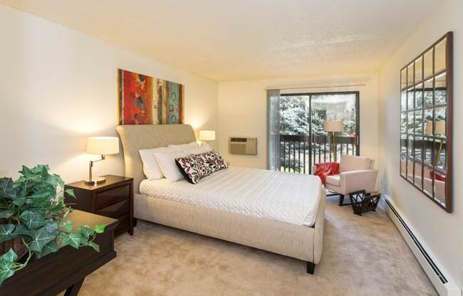 A bright bedroom here at Eagle Ridge featuring a spacious layout with a modern upholstered bed dressed in neutral bedding, a dark wood nightstand with a table lamp, soft carpeted flooring, and a cozy accent chair near a sliding glass door that opens to a private balcony, allowing natural light to fill the room and create a calm, comfortable retreat.