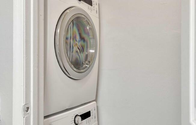 A stacked washer and dryer unit located in a small laundry area against a light gray wall. The washer is on the bottom and the dryer on top, featuring a round glass front on the dryer and control knobs and digital displays on both appliances.