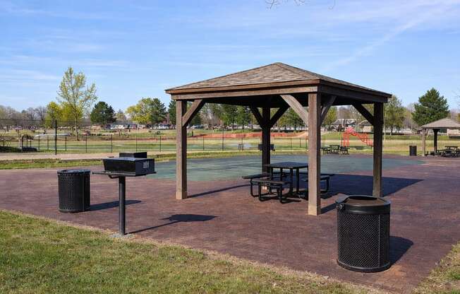 A gazebo sits in the middle of a park with a basketball court in the background.