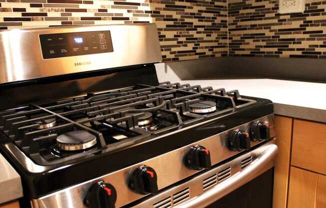 A six burner gas stove in a brookmore apartments kitchen with honey colored wood cabinets and multicolored backsplash.