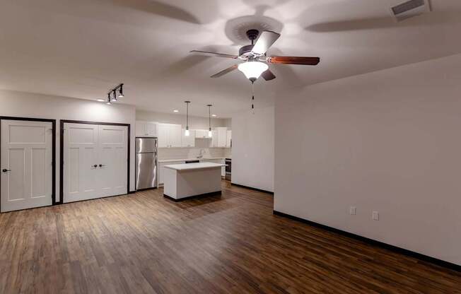 A spacious living room here at The Park featuring warm wood flooring, bright white kitchen with island, ceiling fan, and open layout, creating a modern, inviting, and comfortable living space.