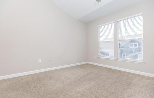 Empty interior room with beige walls and a light carpet. Two windows with white blinds allow natural light to enter. The corner reveals part of a blue building outside, creating a bright and airy atmosphere.