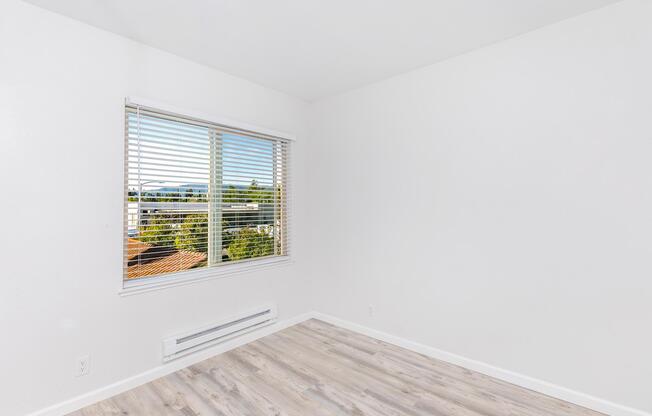 A bright and minimalistic room with white walls and light wooden flooring. A window with blinds offers a view of greenery outside. The space is uncluttered and airy, creating a serene atmosphere.