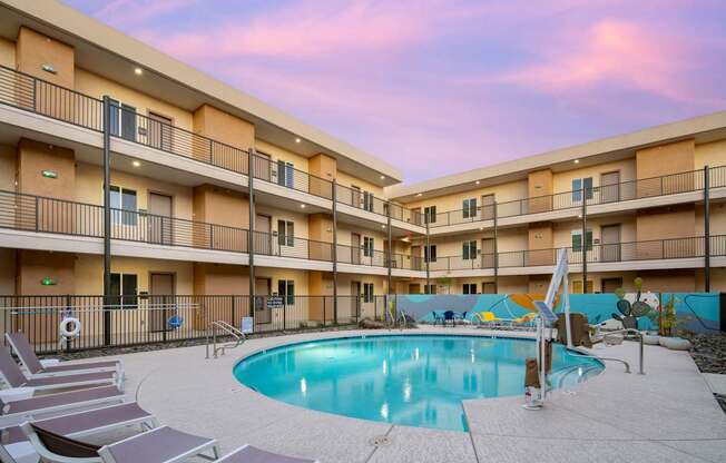 A swimming pool is surrounded by lounge chairs in a courtyard surrounded by apartment buildings.