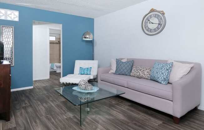 Modern living room in fifteen 50 apartments Las Vegas with grey plank tile floor, grey sofa, glass coffee table, and blue accent wall.