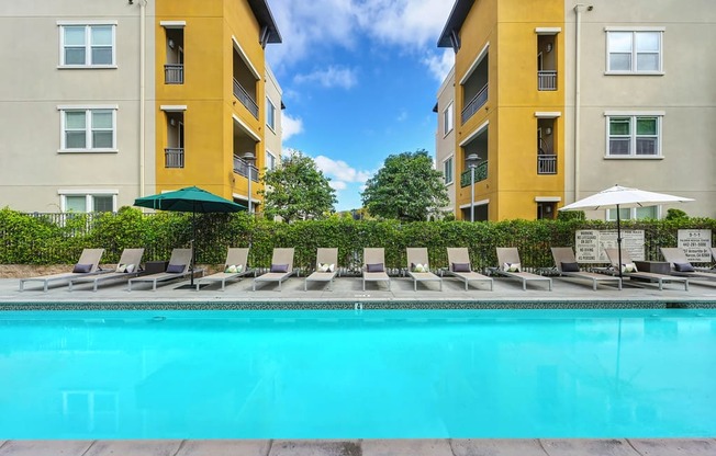 A swimming pool surrounded by lounge chairs and umbrellas in front of apartment buildings.