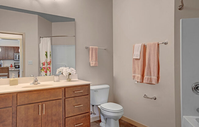 A bright bathroom here at Vue at Pinnacle Park with a wide vanity and mirror, wood cabinets, white countertops, a tub-shower combo with curtain, and warm flooring for a clean, comfortable, and inviting feel.