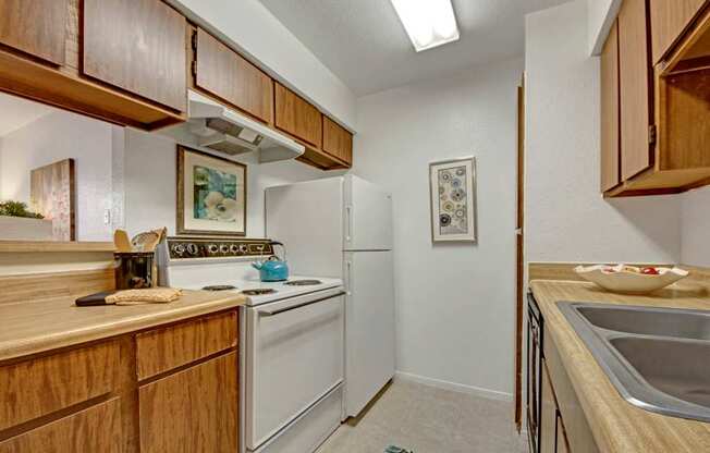 A compact, well-lit kitchen here at Northridge Court featuring warm wood cabinetry, light countertops, a white refrigerator and range, and a double-basin sink, with overhead lighting and neutral finishes that create a clean, functional cooking space.