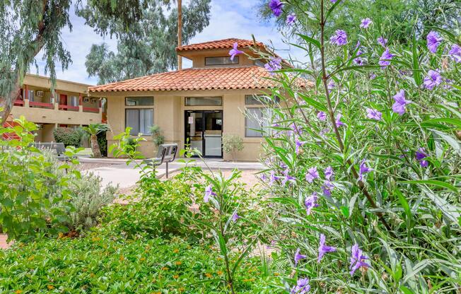 A landscaped area with flowering plants and greenery in the foreground, leading to a building with a red tile roof. The building features large windows and is surrounded by trees. In the background, additional structures are visible, creating a serene outdoor environment.