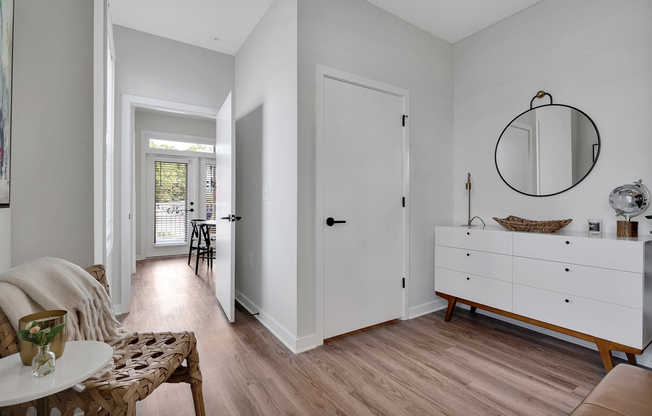 A modern hallway featuring light gray walls and wooden flooring. A round mirror is mounted above a white sideboard with wooden legs, while a wicker chair is positioned nearby. A door leads to another room in the background, showcasing a glimpse of dining furniture through a window.