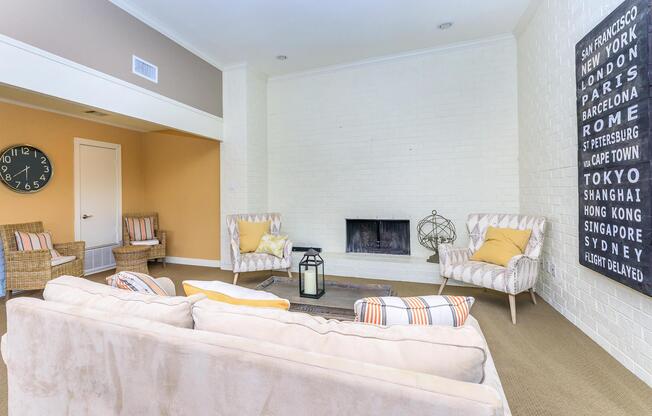 A cozy living room featuring a light-colored sofa with decorative pillows, two armchairs, and a wall-mounted fireplace. To one side, there's a clock and a large board displaying the names of various cities. The room has neutral tones, with a soft carpet and a bright atmosphere.