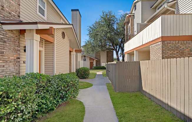 A landscaped pedestrian walkway here at Summertree Place featuring a smooth concrete path winding between apartment buildings with neutral siding, brick accents, privacy fencing, and neatly trimmed greenery, creating a quiet and well-maintained residential setting on a bright day.