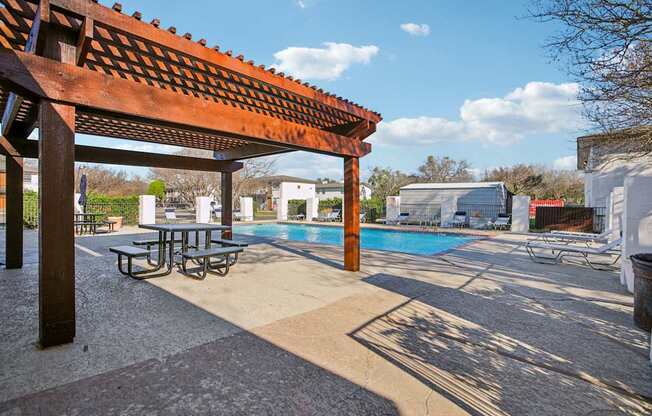 A wooden pergola is over a picnic table next to a pool.