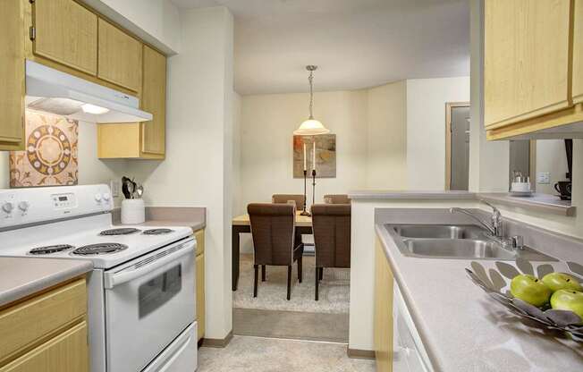 A practical kitchen and dining view here at Allegro showcasing light wood cabinets, neutral countertops, and a full appliance layout with a range, opening to a cozy dining area with a rectangular table, upholstered chairs, and a pendant light, while a breakfast bar and double-basin sink provide a convenient flow for cooking, serving, and everyday meals.