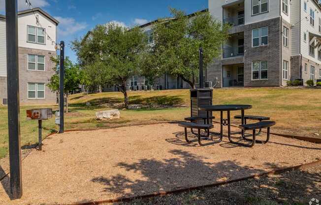 A picnic table sits in the middle of a gravel area with apartment buildings in the background.
