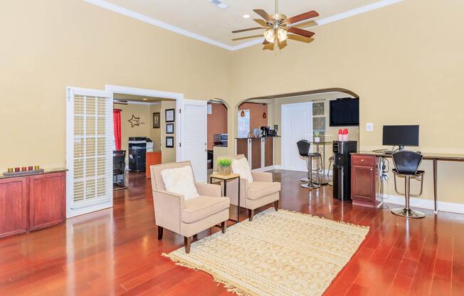 A spacious living room featuring two beige armchairs on a patterned rug. The room has wooden flooring, beige walls, and a ceiling fan. To the left, there's an open archway leading to a kitchen area with dark cabinetry and modern appliances. A small workspace with a computer is visible in the corner.