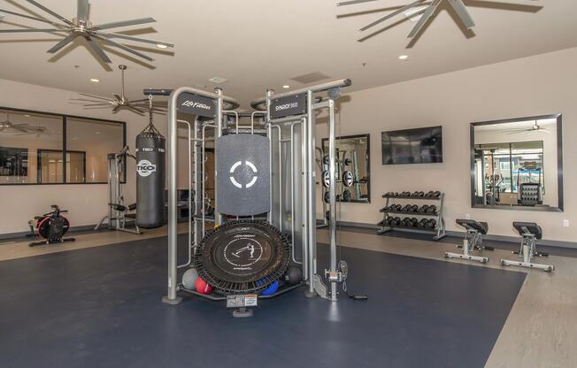 A modern gym interior featuring various fitness equipment, including a multi-station workout machine, free weights, a rowing machine, and a punching bag. The space is well-lit with ceiling fans and mirrors, creating an inviting atmosphere for workouts.