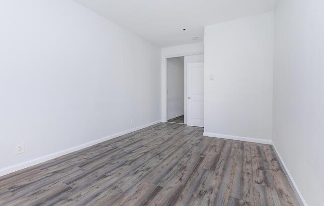 Empty room with light gray walls and wood-like flooring. A door is visible on the left side leading to another room. The space is well-lit, giving a clean and modern appearance, suitable for various uses such as a living area or office.