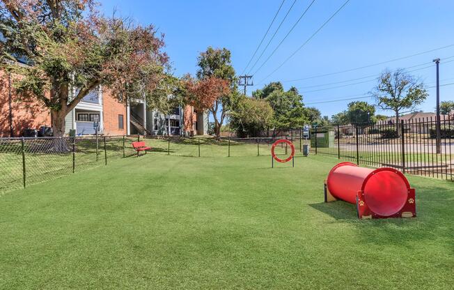 A dog park featuring a grassy area with a red agility tunnel and a hoop. A bench is positioned nearby, and there are trees in the background. The park is enclosed by a fence, with utility poles and buildings visible in the distance under a clear blue sky.