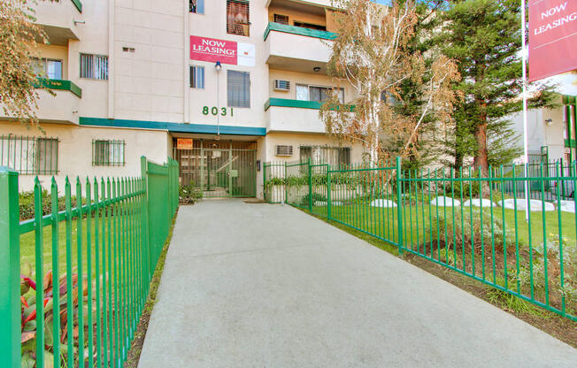 an apartment building with a green fence and a grey walkway leading to the front door