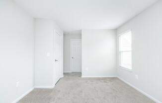 A bright, empty room with light gray walls and beige carpet. There are two closed white doors on the left and a window on the right, allowing natural light to enter. The overall atmosphere is clean and minimalistic, ideal for decorating or furnishing.