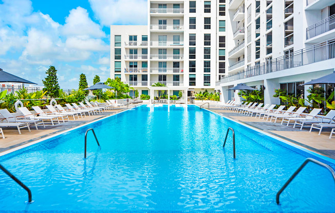 a resort style pool with white chaise lounge chairs and a large white building in the background