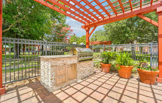 A red pergola is over a stone pillar with potted plants on a brick patio.