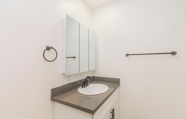 A modern bathroom featuring a white sink with a black faucet, a wall-mounted mirror cabinet, and a simple gray countertop. The walls are painted white, and there is a towel ring and a horizontal towel bar on the walls, creating a clean and minimalist appearance.