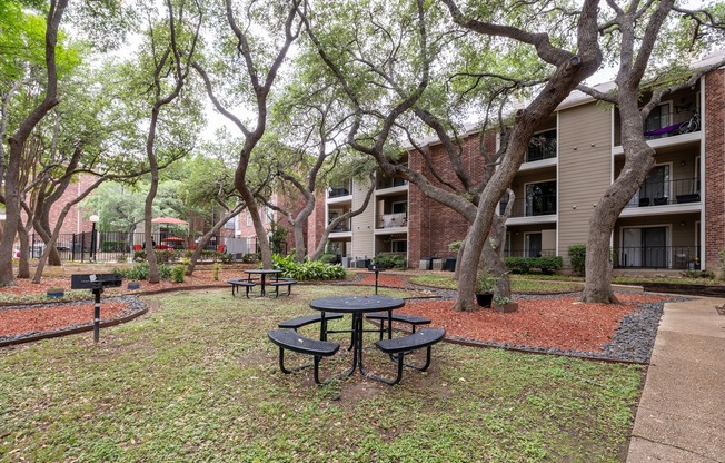 A park area with picnic tables and trees in front of apartment buildings.