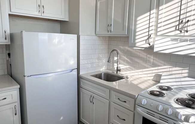 A white refrigerator stands in a kitchen with white cabinets and a white stove.