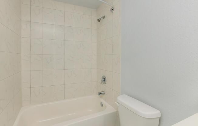 A clean, minimalistic bathroom featuring a beige bathtub with a showerhead. The walls are tiled with light-colored, marbled tiles, and a pale blue wall is visible to the right. A white toilet is positioned beside the bathtub, creating a simple and functional design.
