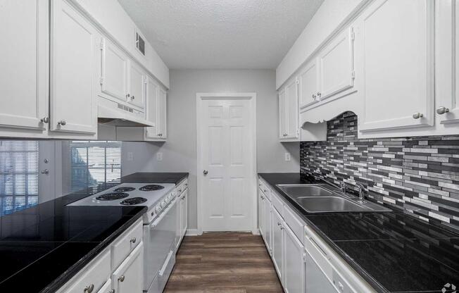 A modern kitchen featuring white cabinets, a black countertop, and a tiled backsplash with gray and black patterns. The design includes a stove with multiple burners, a double sink, and a door leading to another area. The floor is a light wood finish, creating a bright and airy atmosphere.