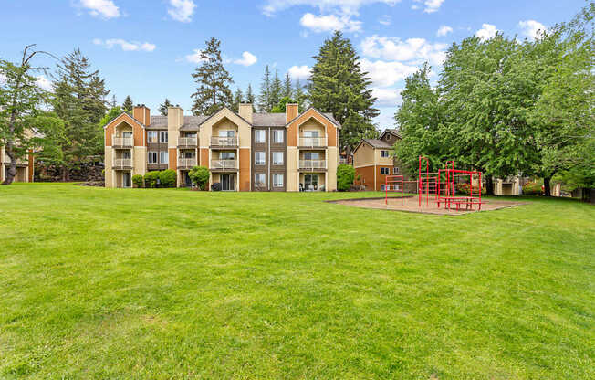 A playground with a red swing set is in the foreground of a grassy area in front of apartment buildings.