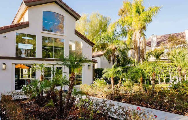 A house with a red tile roof and a white exterior is surrounded by palm trees and other vegetation.