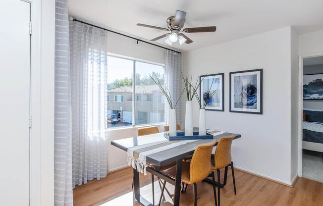 Bright dining area featuring a wooden table with a runner, four yellow chairs, and a large window with sheer curtains. The room is decorated with two framed artworks and a tall white vase with decorative plants. A ceiling fan is visible, and light wood flooring completes the modern look.