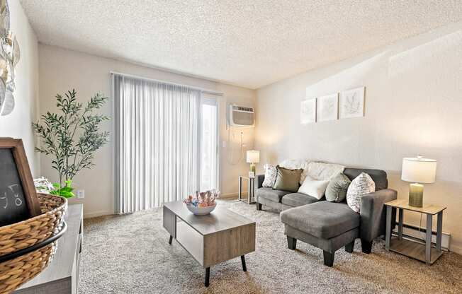 A living room with a grey couch, a coffee table, and a plant A row of apartment buildings with green lawns in front A bedroom with a bed, nightstand, and window at Wildflower, Kennewick, WA 99336.