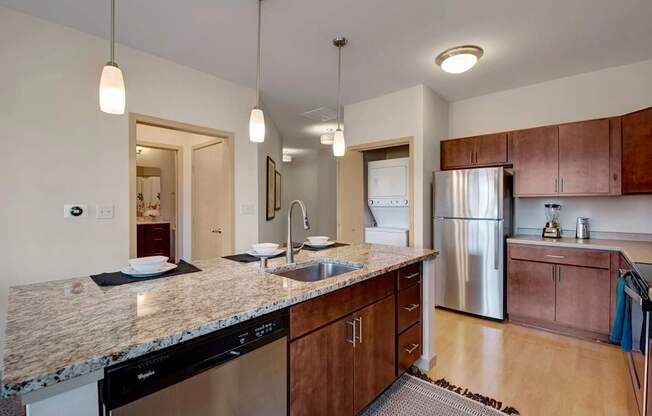 A modern kitchen here at Vue at Pinnacle Park with granite countertops, a large island with sink and bar seating, stainless steel appliances, pendant lighting, and an in-unit stacked washer and dryer nearby for added convenience.
